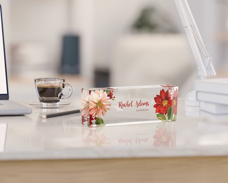 May include: A clear acrylic desk nameplate with a floral design featuring red and white flowers. The nameplate reads "Rachel Adams, Surgeon".
