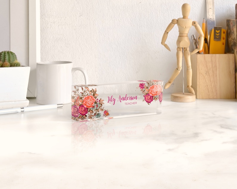 May include: A clear acrylic desk nameplate with a floral design and the text "Lily Anderson Teacher".