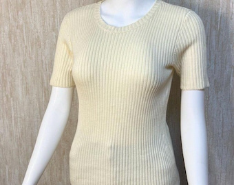 May include: A pale yellow, short-sleeved ribbed knit top with a crew neck, displayed on a mannequin. The top is paired with black trousers. The ribbed texture provides a subtle pattern to the garment.