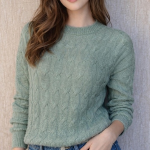 May include: A light green cable-knit sweater with long sleeves. The sweater is paired with blue denim jeans. The model is leaning against a neutral-toned wall. The sweater has a classic, textured design.