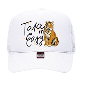 Puede incluir: Gorra de camionero blanca con parte trasera de malla y visera curva. La parte delantera presenta el texto "Take it Easy" en escritura negra, junto con una ilustración de tigre naranja y negro.