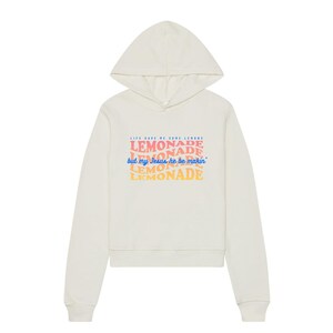 May include: Cream-colored hoodie with a drawstring hood. The front features the text "LIFE GAVE ME SOME LEMONS, LEMONADE, but my Jesus he be makin' LEMONADE" in colorful, layered fonts. A casual, comfortable garment.