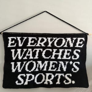 May include: A black and white crocheted wall hanging with the text "EVERYONE WATCHES WOMEN'S SPORTS." The banner is rectangular with a wooden dowel at the bottom and a black string for hanging.