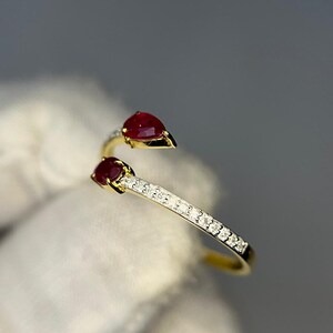 Op de afbeelding: Een goudkleurige ring met twee peervormige, dieprode edelstenen. De ringband is versierd met kleine, heldere edelstenen. De ring is geplaatst tegen een zachte, neutrale achtergrond.