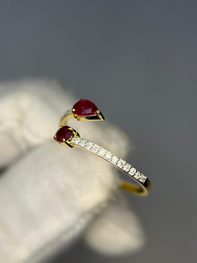 Op de afbeelding: Een goudkleurige ring met twee peervormige, dieprode edelstenen. De ringband is versierd met kleine, heldere edelstenen. De ring is geplaatst tegen een zachte, neutrale achtergrond.