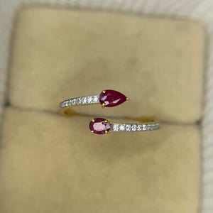 Op de afbeelding: Een goudkleurige ring met twee peervormige, dieprode edelstenen. De ringband is versierd met kleine, sprankelende witte stenen. Het ontwerp is open, met de edelstenen aan weerszijden van de band.