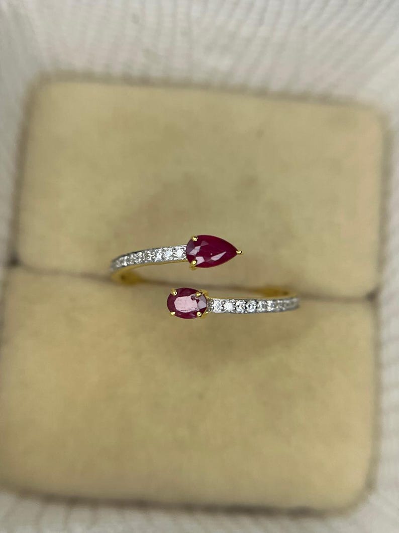 Op de afbeelding: Een goudkleurige ring met twee peervormige, dieprode edelstenen. De ringband is versierd met kleine, sprankelende witte stenen. Het ontwerp is open, met de edelstenen aan weerszijden van de band.