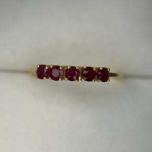Op de afbeelding: Een gouden ring met vijf ronde, dieprode robijnen in een horizontale rij. Elke robijn is vastgezet met kleine gouden pootjes, waardoor een doorlopende lijn van edelstenen over de bovenkant van de ring ontstaat met een klassiek ontwerp.
