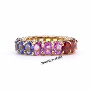 Könnte beinhalten: Ein goldener Eternity-Ring mit einer durchgehenden Reihe oval geschliffener Edelsteine in einem Regenbogen-Farbverlauf aus Blau, Violett, Pink und Rot. Der Text JewelsLoveGifts ist in Schwarz unter dem Ring auf weißem Grund gedruckt.