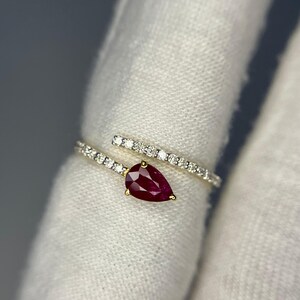 Op de afbeelding: Een gouden ring met een peervormige, dieprode edelsteen als middelpunt. De band van de ring heeft een spiraalvormig ontwerp, met kleine, heldere edelstenen langs de band. De ring is geplaatst tegen een witte achtergrond.