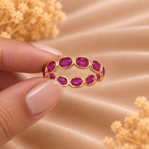 Puede incluir: Un anillo de banda dorada que presenta una fila continua de piedras preciosas ovaladas y facetadas de color rosa magenta, engastadas en biseles de metal individuales. El anillo muestra un diseño uniforme con metal dorado pulido.