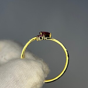 Op de afbeelding: Een gouden ring met twee donkerrode edelstenen. De ring heeft een delicate band en een kleine, vierkante edelsteen naast een grotere, peervormige edelsteen. De ring wordt tegen een neutrale achtergrond gehouden.