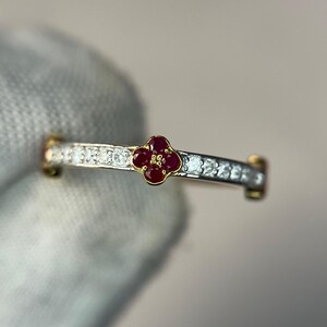 Op de afbeelding: Een ring met een centraal vierbladig bloemmotief, bezet met vier kleine rode robijnen in een geelgouden zetting. De ringband is gemaakt van zilverkleurig metaal en is aan weerszijden versierd met een rij kleine, heldere, ronde witte diamanten.