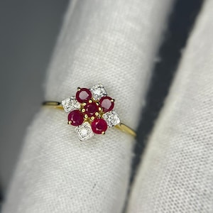 May include: A gold ring featuring a floral design with red gemstones and small white stones. The ring's band is thin and gold, and the gemstones are arranged in a cluster. The ring is set against a white background.