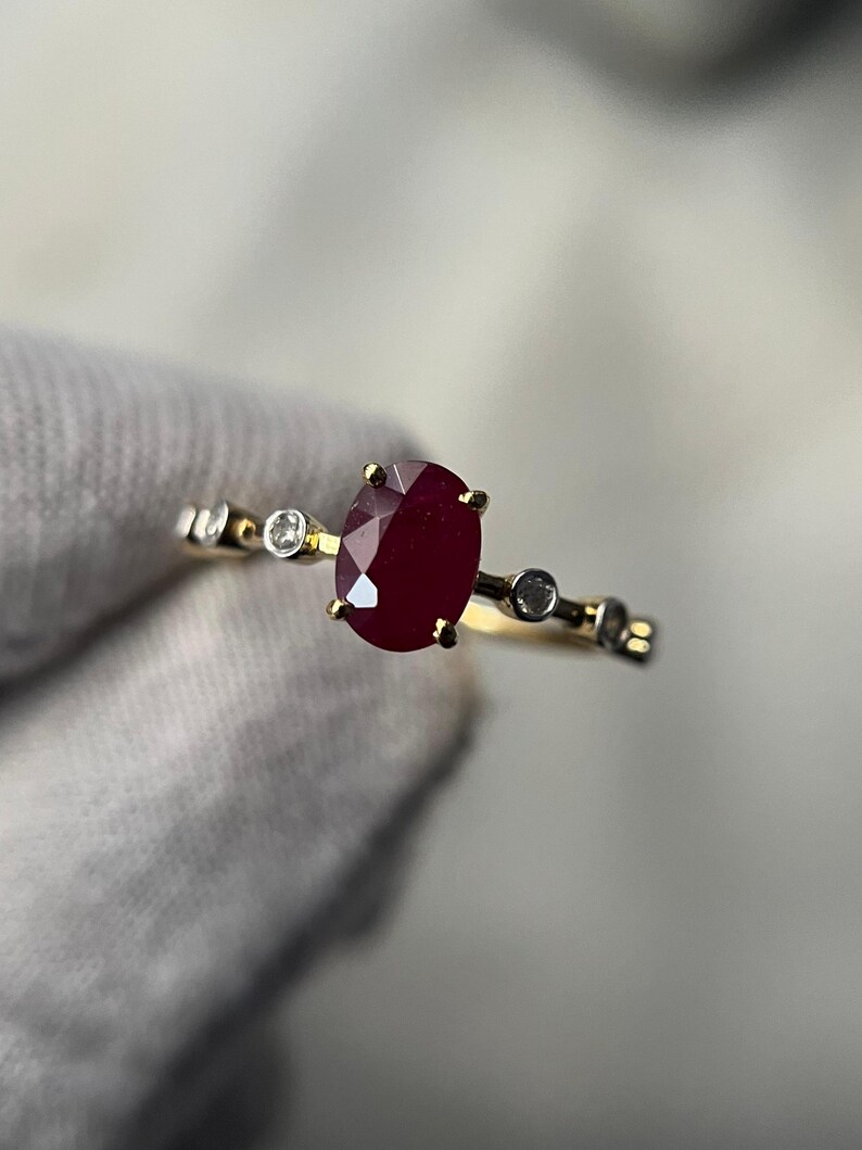 May include: A gold ring featuring a large, oval-cut ruby as the primary stone. The ring is accented with small, round diamonds and has a delicate band. The ruby is a deep red color, and the diamonds sparkle.