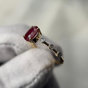 May include: A gold ring featuring a large, oval-cut ruby gemstone as the centerpiece. The ring is accented with three smaller, round diamonds set along the band. The ruby is a deep red color, and the ring is set against a neutral background.