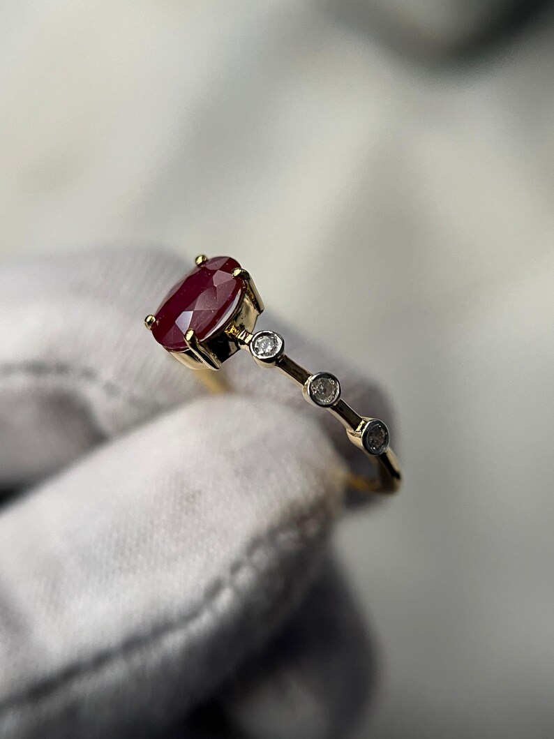 May include: A gold ring featuring a large, oval-cut ruby gemstone as the centerpiece. The ring is accented with three smaller, round diamonds set along the band. The ruby is a deep red color, and the ring is set against a neutral background.