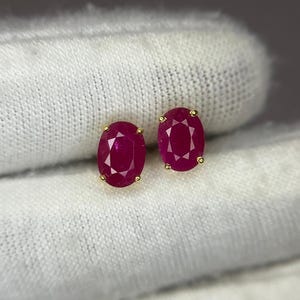 May include: A pair of oval-cut, deep red ruby stud earrings set in yellow gold prongs. Each gemstone features a faceted surface that reflects light, showcasing a vibrant crimson hue. The earrings are positioned side by side, highlighting the symmetrical oval shape and the polished gold metal settings.