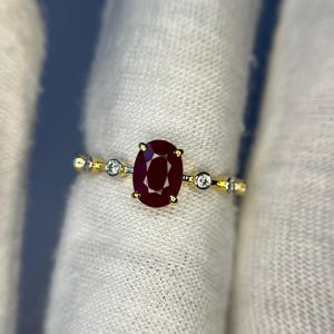 May include: A gold ring featuring a large, oval-cut, deep red ruby as the centerpiece. The band is adorned with small, round, clear gemstones and delicate gold accents, creating an elegant and refined design.