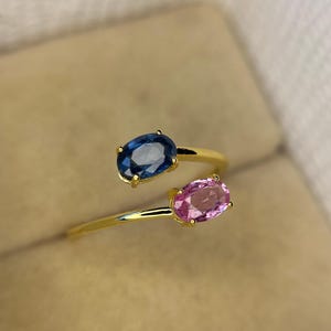 May include: A gold-colored ring featuring two oval gemstones, one blue and one pink. The gemstones are set in a bypass design, with the band curving around the finger. The ring is set against a neutral background.