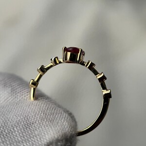 May include: A gold ring with a small, round, red gemstone at the center. The band features a series of small, rectangular accents. The ring is held by a hand wearing a white glove.