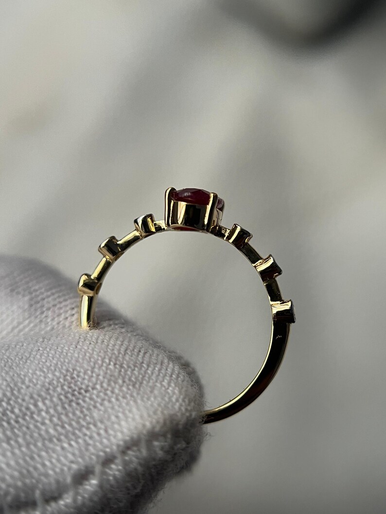 May include: A gold ring with a small, round, red gemstone at the center. The band features a series of small, rectangular accents. The ring is held by a hand wearing a white glove.