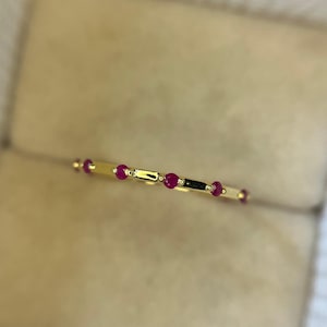 Peut inclure: Une fine bague en or ornée d'une série de petites pierres précieuses rouges vives. Le design de la bague intègre une disposition linéaire des pierres rouges, entrecoupées de segments dorés. La bague est présentée sur un fond neutre et doux.