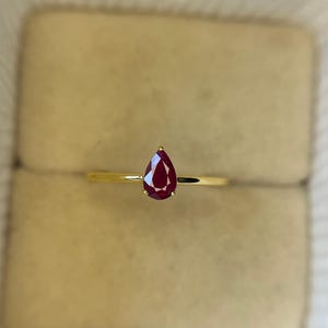 May include: A gold ring with a teardrop-shaped, deep red gemstone. The ring has a slender gold band, and the gemstone is the main feature, set against a neutral backdrop. This ring is likely a piece of jewellery.