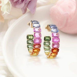 May include: Two gold hoop earrings, each featuring a spectrum of rectangular gemstones. The colours transition from green to yellow, orange, red, pink, and blue. The earrings are presented on a white surface.