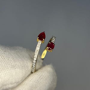 Op de afbeelding: Een goudkleurige ring met twee rode edelstenen. E&eacute;n is peervormig, de andere ovaal. De band is bezet met kleine, heldere edelstenen. De ring wordt getoond tegen een grijze achtergrond.
