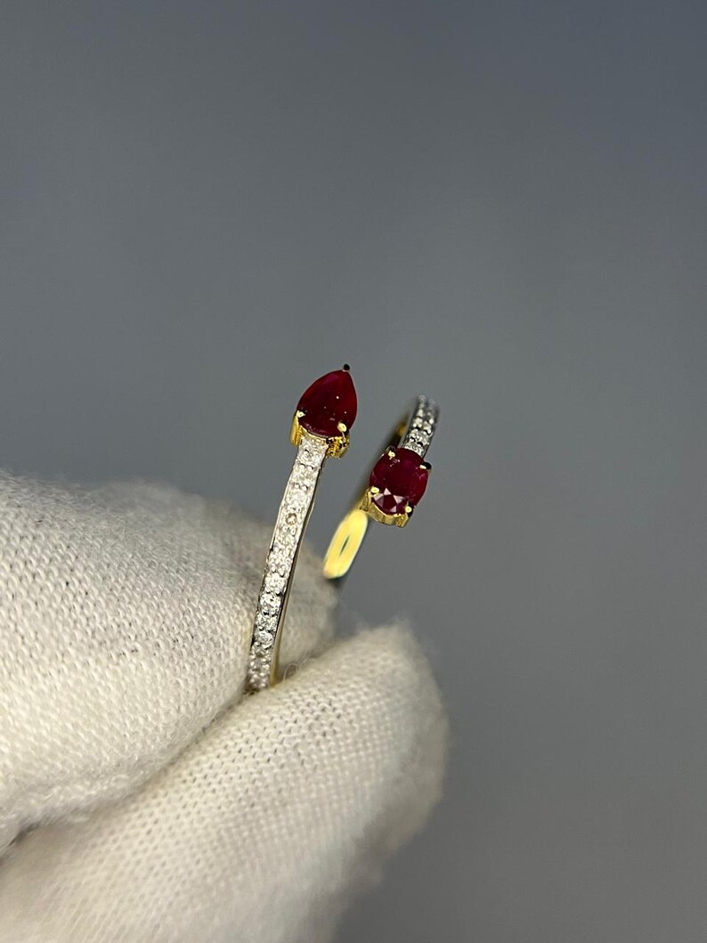 Op de afbeelding: Een goudkleurige ring met twee rode edelstenen. E&eacute;n is peervormig, de andere ovaal. De band is bezet met kleine, heldere edelstenen. De ring wordt getoond tegen een grijze achtergrond.