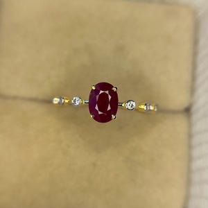 May include: A gold ring featuring a large oval-cut ruby center stone, flanked by two smaller round diamonds. The ring's band is a thin, polished gold, with small accent stones. The ruby is a deep, rich red color.
