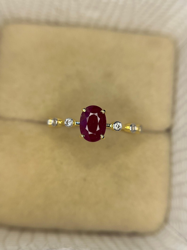 May include: A gold ring featuring a large oval-cut ruby center stone, flanked by two smaller round diamonds. The ring's band is a thin, polished gold, with small accent stones. The ruby is a deep, rich red color.
