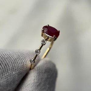 May include: A gold ring featuring a large oval ruby gemstone and three smaller round diamonds. The ruby is a deep red color, and the diamonds sparkle. The ring has a delicate, minimalist design.