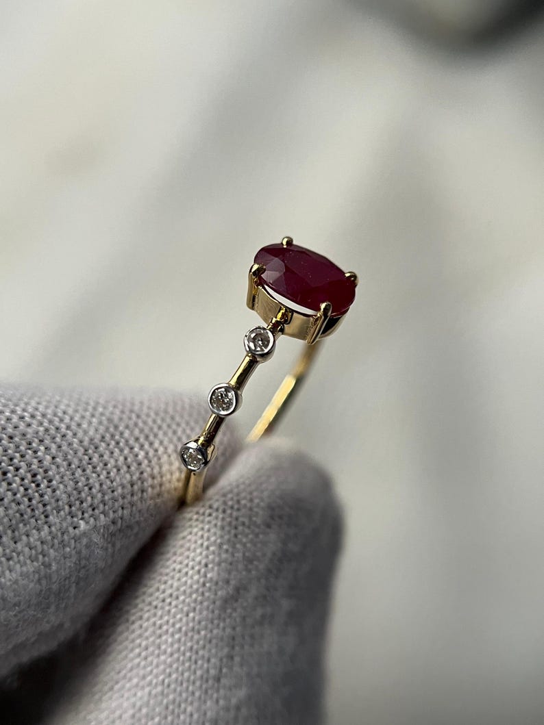 May include: A gold ring featuring a large oval ruby gemstone and three smaller round diamonds. The ruby is a deep red color, and the diamonds sparkle. The ring has a delicate, minimalist design.