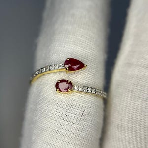 Op de afbeelding: Een goudkleurige ring met twee rode edelstenen, een peervormige en een ovale, gezet op banden met kleine, heldere stenen. De ring heeft een open ontwerp, met de edelstenen aan weerszijden.