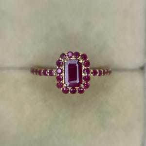 May include: A gold ring featuring a central emerald-cut ruby surrounded by a halo of smaller round rubies. The band is also set with a series of round rubies extending along the sides, creating a continuous display of deep red gemstones.