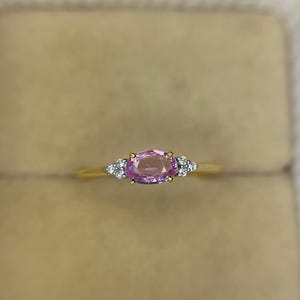 May include: A gold ring featuring a large oval pink gemstone, flanked by smaller white gemstones on each side. The ring is set against a neutral background, highlighting the jewelry's design and color.