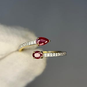 Op de afbeelding: Een goudkleurige ring met twee rode edelstenen, een peervormige en een ovale. De ringband is bezet met kleine, witte stenen. Het ontwerp is open, met de edelstenen aan weerszijden van de vinger. Een elegant sieraad.