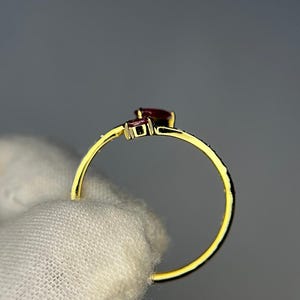 Op de afbeelding: Een gouden ring met twee donkerrode edelstenen. De ring heeft een delicate band en een kleine, vierkante steen naast een grotere, peervormige steen. De ring wordt tegen een neutrale achtergrond gehouden.