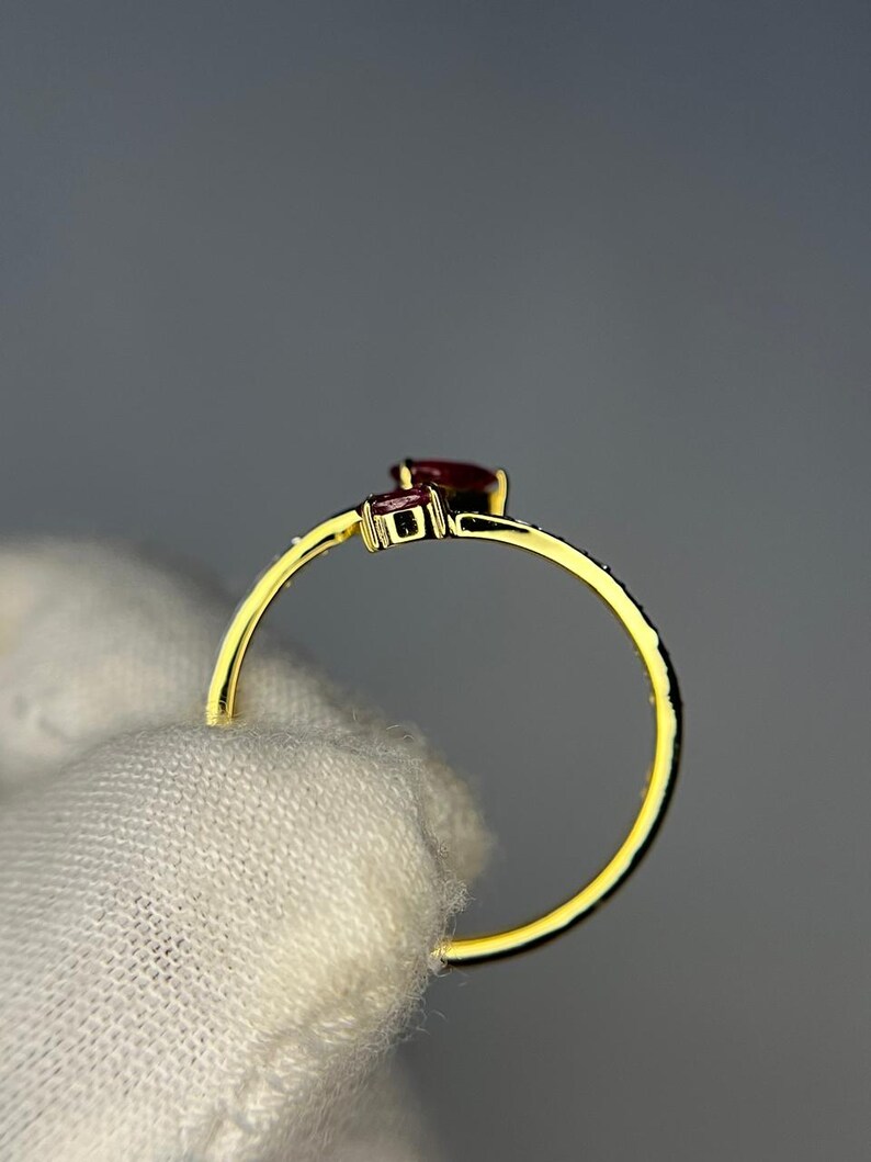 Op de afbeelding: Een gouden ring met twee donkerrode edelstenen. De ring heeft een delicate band en een kleine, vierkante steen naast een grotere, peervormige steen. De ring wordt tegen een neutrale achtergrond gehouden.