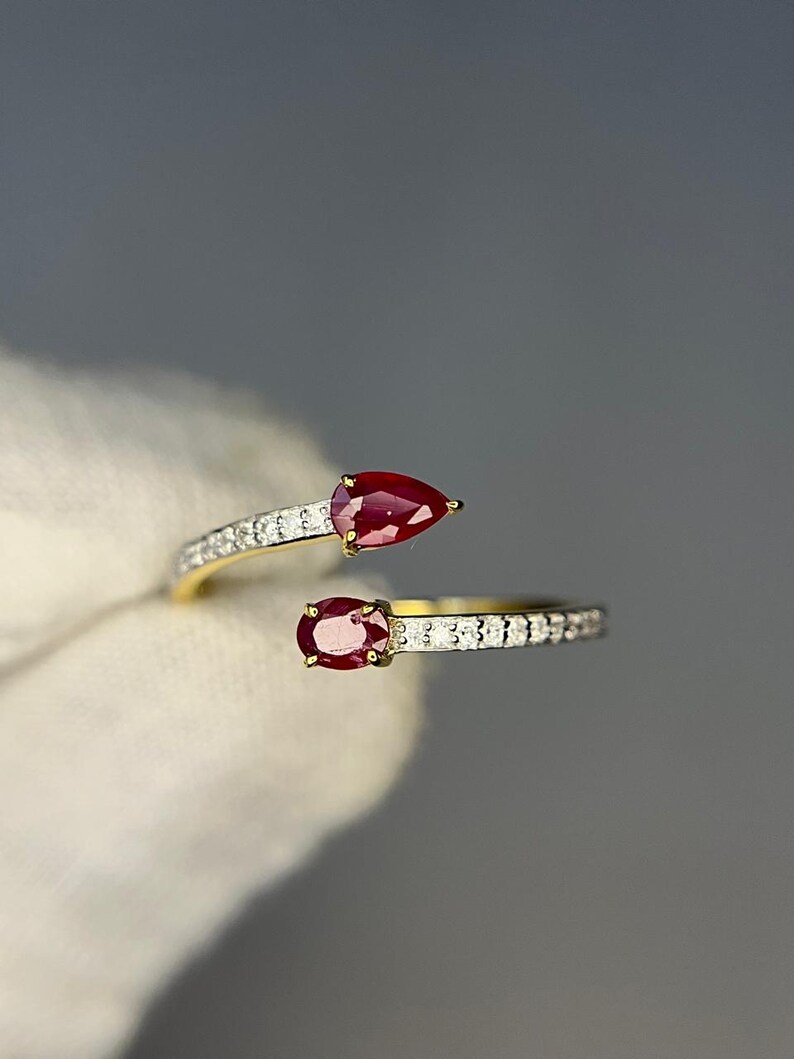 Op de afbeelding: Een goudkleurige ring met twee levendige rode edelstenen, een peervormige en een ovale. De ringband is versierd met kleine, sprankelende witte stenen. Het open ontwerp heeft de edelstenen aan weerszijden van de vinger.