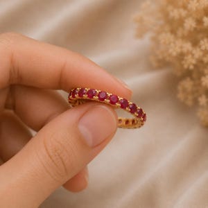 Könnte beinhalten: Ein goldener Eternity-Ring mit einer durchgehenden Reihe runder, leuchtend roter Edelsteine in einer polierten Metallfassung. Der Ring zeigt ein einheitliches Design, bei dem jeder Stein sicher von kleinen Krappen gehalten wird.
