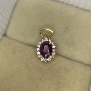 May include: A gold pendant featuring a central oval-cut deep red ruby surrounded by a halo of small, round, clear white stones. The pendant includes a gold bail at the top for attachment to a chain, showcasing a classic cluster design.