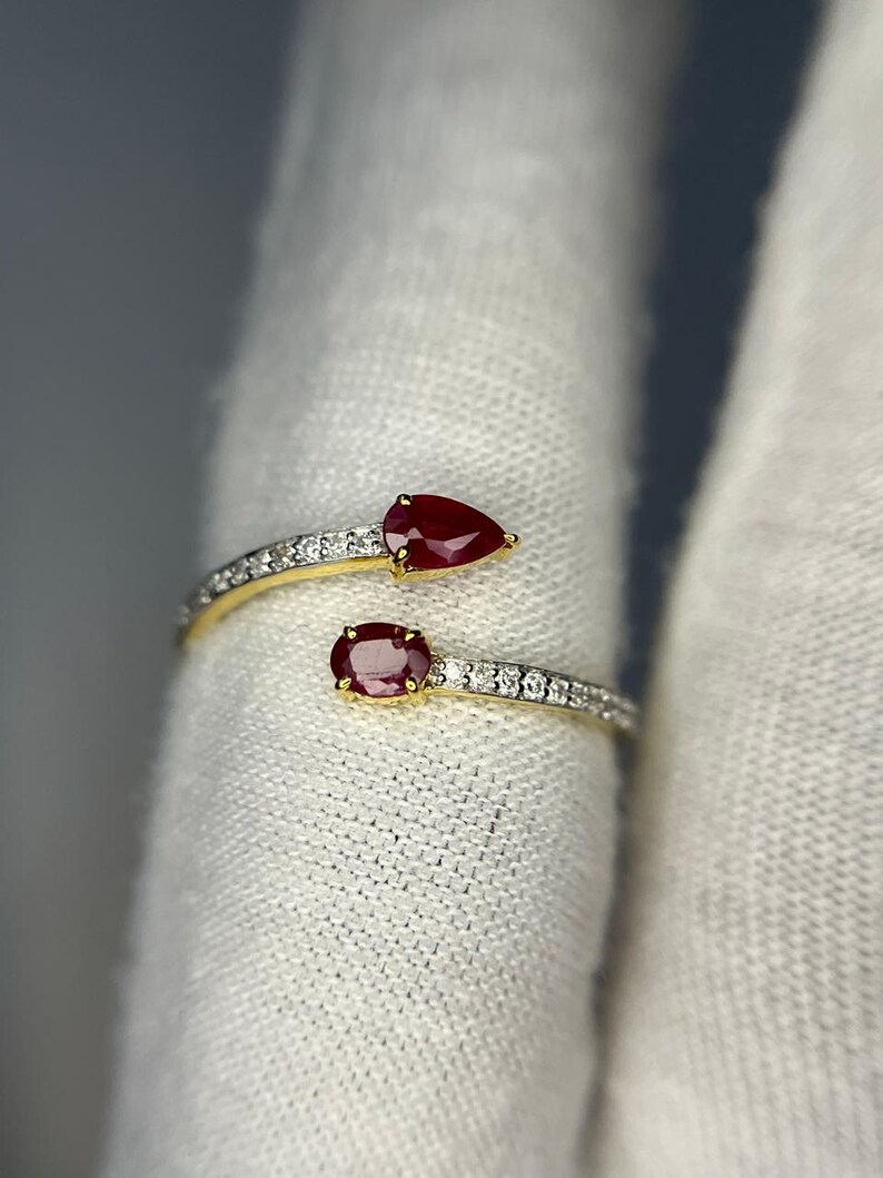 Op de afbeelding: Een goudkleurige ring met twee rode edelstenen, een peervormige en een ovale, gezet op banden met kleine, heldere stenen. De ring heeft een open ontwerp, met de edelstenen aan weerszijden.