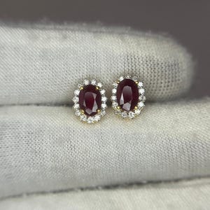 May include: A pair of stud earrings featuring oval-cut deep red rubies at the centre, each surrounded by a halo of small, round, clear white diamonds set in a yellow gold metal frame, creating a classic floral-inspired cluster design.