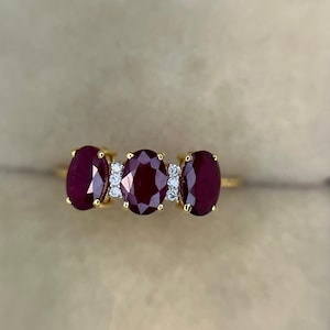 Puede incluir: Un anillo de oro con tres rubíes de color rojo intenso y corte ovalado dispuestos horizontalmente. Los rubíes están separados por dos pequeñas secciones verticales, cada una con tres diminutos diamantes blancos redondos en engastes de oro.