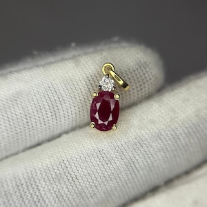 May include: A gold pendant featuring an oval-cut deep red ruby gemstone set in four prongs, topped with a single round brilliant-cut white diamond in a silver-toned setting, complete with a gold bail for attachment to a necklace chain.