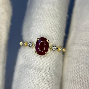 May include: A gold ring featuring a large, oval-cut ruby gemstone as the centerpiece. The ruby is a deep, rich red color. The ring also has small, round, clear gemstones on either side of the ruby, adding sparkle.