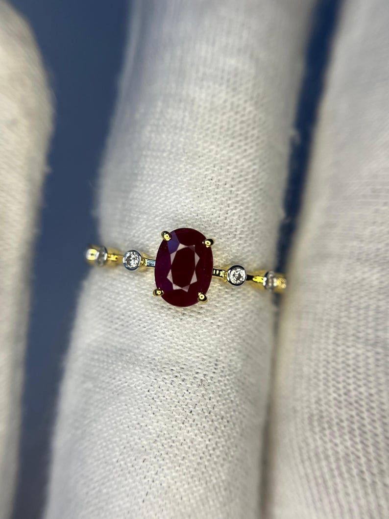 May include: A gold ring featuring a large, oval-cut ruby gemstone as the centerpiece. The ruby is a deep, rich red color. The ring also has small, round, clear gemstones on either side of the ruby, adding sparkle.
